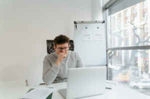 A man in grey sweaters thinking while sitting in front of a laptop and documents inside an office