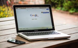 A macbook with google search on screen and phone on a wooden table.