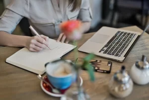 A person writing on a notebook beside a laptop